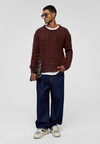 Bruine kabelgebreide trui met een ronde halslijn, gedragen over een witte blouse. Gecombineerd met losse blauwe jeans en witte sneakers, met een donkere clutch in de hand.
