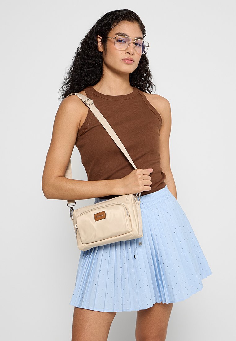 Beige Umhängetasche aus weichem Material, mit einem Reißverschlussverschluss, einer frontalen Tasche mit einem braunen Etikett und einem verstellbaren Trageriemen.