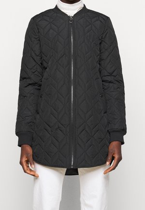 Personne portant une veste matelassée noire avec une fermeture éclair frontale par-dessus un col roulé blanc et un pantalon blanc, debout devant un fond uni.