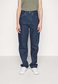 Jeans de mezclilla azul oscuro con cintura alta, corte holgado y costuras visibles. Combinados con una blusa verde clara y zapatillas blancas.