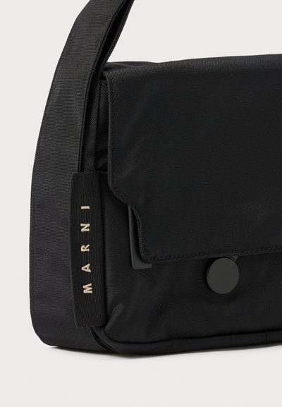 Melnas neilona crossbody soma ar atloku, kas satur spiedogu, logotipa birku un pārklātu tekstūru. Kompakta un taisnstūrveida dizaina.