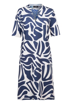 Vrouw draagt een witte knielange jurk met een abstract marineblauw patroon, V-hals, korte mouwen en witte sandalen met hakken, staande tegen een witte achtergrond.