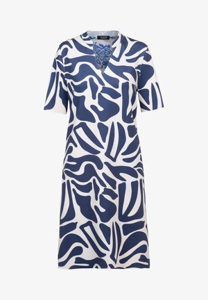 Femme portant une robe blanche mi-longue avec un motif abstrait bleu marine, col en V, manches courtes et sandales blanches à talons, debout devant un fond blanc.
