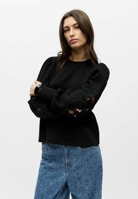 Svart genser med pufferm og blomsterbroderier og utskjæringer. Kombinert med teksturerte blå denimjeans. Enkelt hvitt bakgrunn.