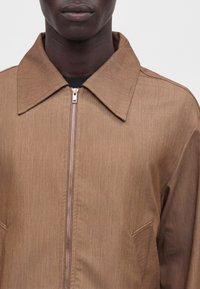 Homme portant une veste marron zippée avec un col large et des détails de coutures visibles, sur un fond blanc uni.