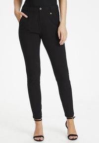 Pantalon noir slim fit avec une taille plate, poches latérales et un petit emblème doré près de la taille. Fabriqué à partir d'un matériau lisse et extensible.