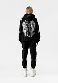 Persona de espaldas vistiendo sudadera con capucha y pantalones negros con patrones de plumas en forma de alas en blanco y plata, y botas blancas con cordones sobre fondo blanco.