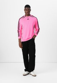 Pitkähihaisen kirkkaan pinkin urheilupaidan mustilla raidoilla, harmailla yksityiskohdilla ja adidas-logolla, yhdistetty mustiin housuihin ja valkoisiin kenkiin.