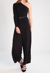 Top nero a spalla unica con texture a coste, abbinato a pantaloni neri larghi e morbidi. Una cintura nera sottile segna il punto vita. Si indossano tacchi neri.