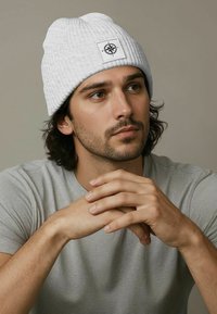 White ribbed beanie with a textured finish, featuring a square patch with a compass design, worn by a person with medium-length dark hair.