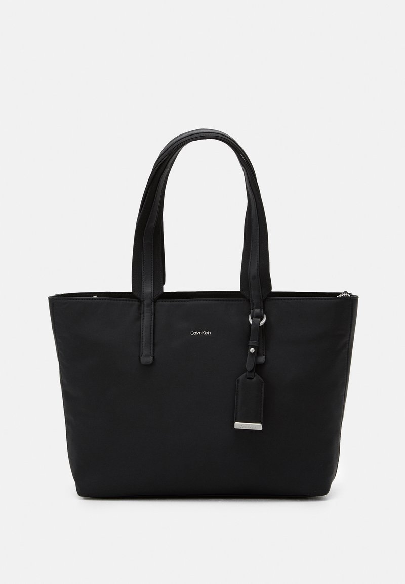 Sac cabas rectangulaire noir avec double bandoulière, petit logo Calvin Klein et étiquette suspendue avec détail métallique.