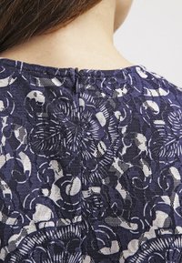 Gros plan sur le haut du dos d'une personne portant un haut en dentelle à motif bleu marine et blanc avec une petite fermeture éclair au niveau de l'encolure.