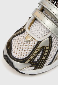 Αθλητικό παπούτσι με άνω μέρος από δίχτυ σε λευκό, μαύρο και ασημί αποχρώσεις. Διαθέτει τρεις μεταλλικές λουρίδες, υφασμένο πάτο και διακριτική λεπτομέρεια λογότυπου.