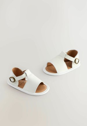 Sandales en cuir blanc avec orteils ouverts et sangle réglable, dotées d'une boucle circulaire en métal doré. Semelle intérieure en brun doux et semelle blanche.