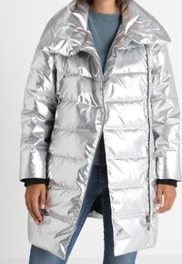 Srebrna, metaliczna kurtka puchowa ze dużym kołnierzem, zapinana na zamek z przodu, pikowana. Posiada długie rękawy i długość do połowy uda.