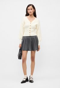 Cardigan crem cu tricotaj tip cablu, cu mâneci lungi și pufoase, cu nasturi aurii, asociat cu o fustă gri plisată, o geantă neagră și pantofi Mary Jane negri.