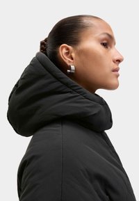 Chaqueta negra acolchada con una gran capucha plegada. Textura suave y diseño minimalista. Pendientes circulares plateados acentúan el look.