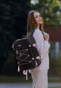 Tmavě hnědý batoh s růžovými akcenty, má obdélníkový tvar, více přihrádek a detail v podobě elastického šňůrování. Nastavitelné ramenní popruhy.