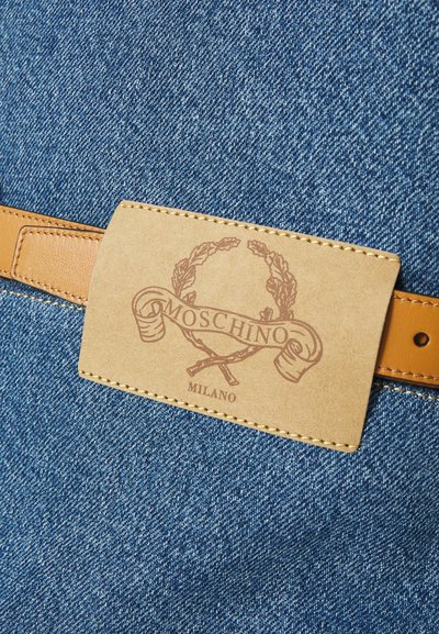 Denim fabric with a tan leather tag. The tag features a printed logo in brown, bordered by a decorative pattern and stitching.