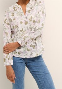 Langærmet bluse i hvidt stof med pastelfarvede blomsterdesigns i pink og grøn. Har en V-udskæring og løs pasform. Sammen med blå jeans.