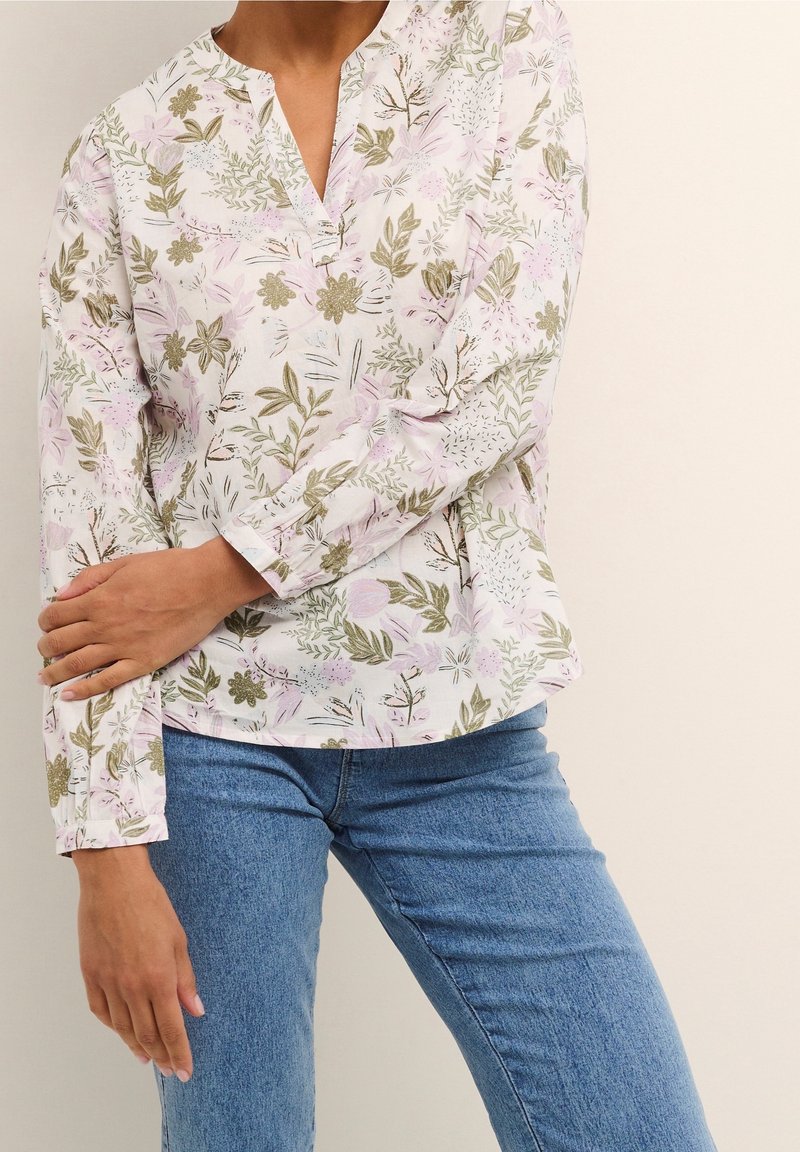 Langærmet bluse i hvidt stof med pastelfarvede blomsterdesigns i pink og grøn. Har en V-udskæring og løs pasform. Sammen med blå jeans.