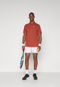 Červené polo tričko s límečkem a logem Nike, bílé šortky, černé sportovní boty a tenisová raketa v jedné ruce, držící tenisový míček.