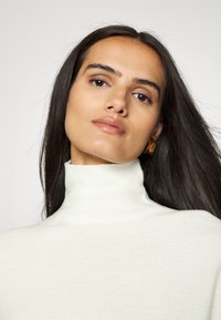 Cremfarbener Rollkragenpullover aus weichem Strickmaterial, mit hohem Hals und einer glatten Textur. Das Modell trägt goldene Ohrringe und hat dunkles, fließendes Haar.