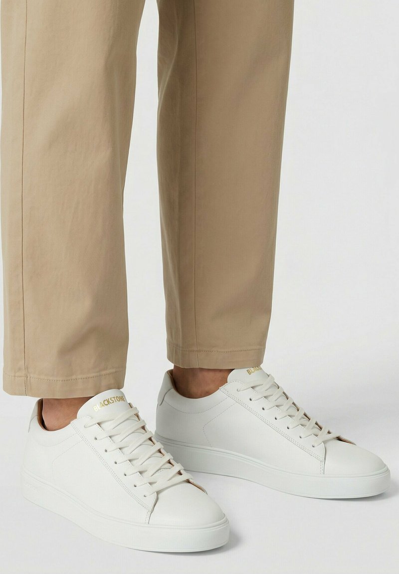 Persoon draagt beige rechte broekspijpen en witte lage veterneakers met "BLACKSTONE"-branding op de tong.