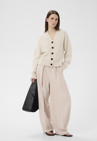 Frau trägt eine beige, geknöpfte Strickjacke, weite, hellrosa Hosen, schwarze Sandalen, hält eine schwarze Handtasche und steht vor einem schlichten weißen Hintergrund.