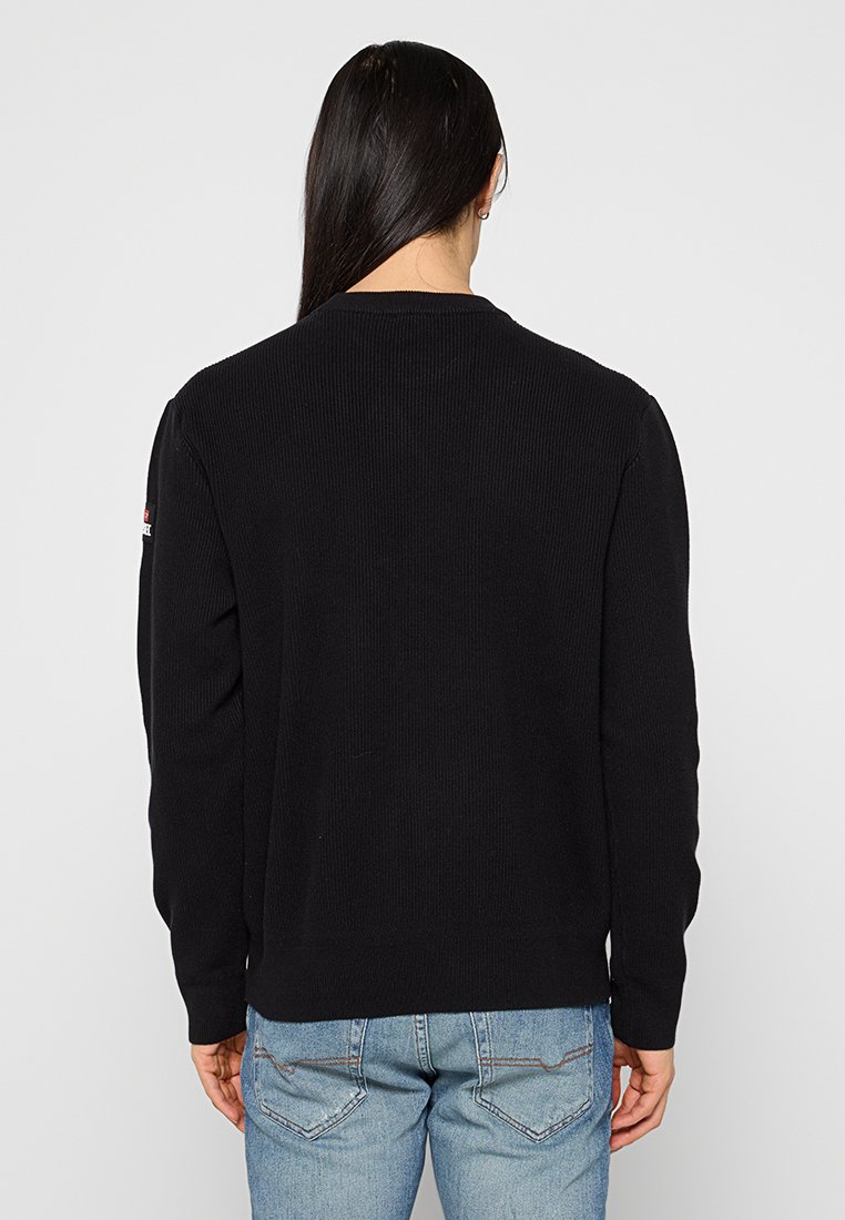 Persona con lunghi capelli scuri che indossa un maglione nero e jeans blu, girata di spalle rispetto alla fotocamera su uno sfondo chiaro e semplice.