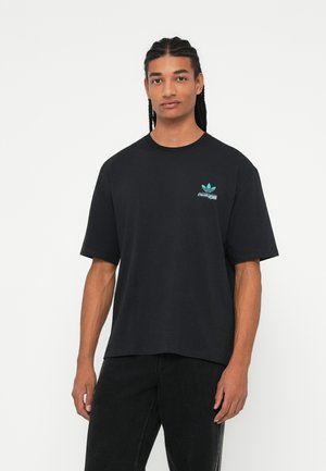 Ung mand med fletninger iført en sort Adidas t-shirt med et lille blåt logo, stående foran en ensfarvet hvid baggrund.