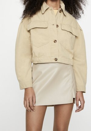 Femme portant une veste courte beige à gros boutons, sur une mini jupe en similicuir beige clair avec une fente sur le côté.