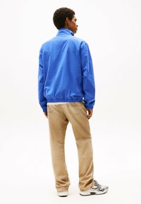 Giacca antivento blu con colletto alto e vita elastica, indossata sopra pantaloni beige dal taglio largo e sneaker grigie.