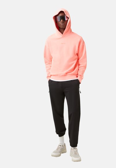 Różowy rozpinany sweatshirt z logo z przodu, w połączeniu z czarnymi zwężanymi spodniami i beżowymi trampkami; model nosi ciemne okulary przeciwsłoneczne.