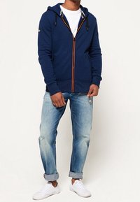 Hombre vistiendo una sudadera con cremallera azul marino, camiseta blanca, jeans claros arremangados y zapatillas blancas, de pie contra un fondo liso.