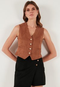 Vestă maro din piele întoarsă cu decolteu în V, cinci nasturi din lemn și două buzunare frontale, asortată cu pantaloni scurți negri cu un detaliu similar de nasturi.