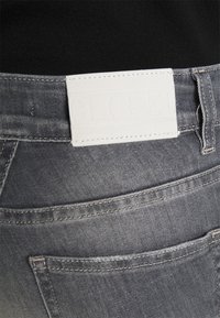 Szare dżinsy z denimu z białą metką z tkaniny, na której znajduje się wytłoczone hasło "CLOSED". Widoczne przeszycia i tylne kieszenie. Tekstura wydaje się miękka i elastyczna.