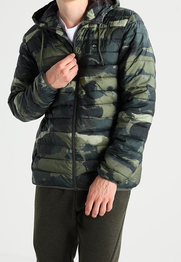 Homme portant une doudoune à motif camouflage vert et noir avec une capuche, zippée à moitié, debout devant un fond uni.