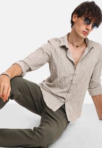Camicia grigia chiaro con texture, colletto, chiusura a bottoni e maniche arrotolate, abbinata a pantaloni verde scuro. Nessun motivo o accento visibile.