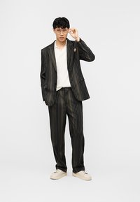 Νέος άνδρας που διορθώνει τα γυαλιά του, φοράει μαύρο κοστούμι με λεπτές ρίγες και χρυσές λεπτομέρειες, λευκό πουκάμισο και κρεμ αθλητικά παπούτσια, στέκεται σε απλό φόντο.