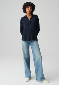 Camicia button-up blu navy con colletto, abbinata a jeans azzurri a gamba larga e sneakers bianche, su uno sfondo chiaro e uniforme.