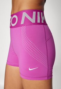 Rausvos sportinės šortai su plačiu pilku Nike logotipo juosmeniu, su iškilniais taškų raštais šone ir mažuoju baltuoju swoosh logotipu.