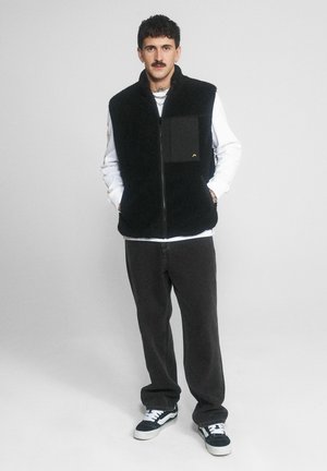 Mężczyzna w czarnej polarowej kamizelce na białej koszuli z długim rękawem, ciemnych luźnych spodniach, czarno-białych sportowych butach, stoi z rękami w kieszeniach.