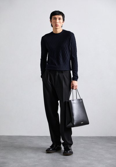 Schwarzer Strickpullover mit texturiertem Muster, maßgeschneiderte schwarze Hose und schwarze Leder-Tote. Das Model trägt glänzende schwarze Schuhe.
