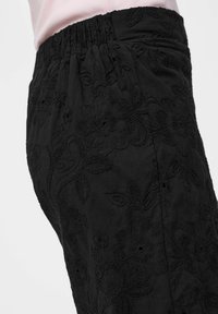 Gros plan d'un pantalon noir avec une ceinture élastique présentant une broderie florale complexe et un haut rose pâle rentré à l'intérieur.