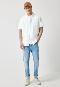 Witte kortarmige shirt, lichte denim jeans, tan suède pet en witte sneakers, die een casual zomerstijl tonen.