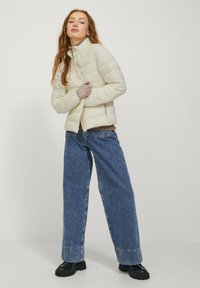 Lichtbeige pufferjas met horizontale gestikte ontwerp, hoge kraag en ritssluiting, gecombineerd met wijde blauwe denim jeans en zwarte laarzen.