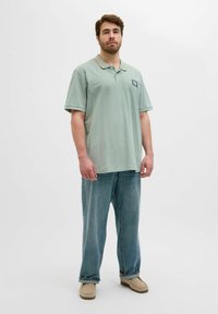 Hele roheline polo särk, millel on oranž krae ja logo pealdis, koos laia lõikega siniste teksade ja beežide slip-on kingadega.