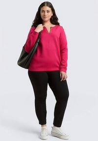 Fuchsiafarbenes Sweatshirt mit V-Ausschnitt und strukturierten Akzenten, kombiniert mit schwarzen, enganliegenden Hosen und weißen Turnschuhen, gehalten mit einer schwarzen Tragetasche.