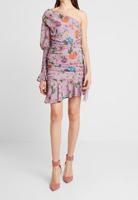 Žena v lila květinových šatech s jedním ramienkem a volánkovým lemem, doplněná o růžové sandály na kotníku, na jednobarevném bílém pozadí.
